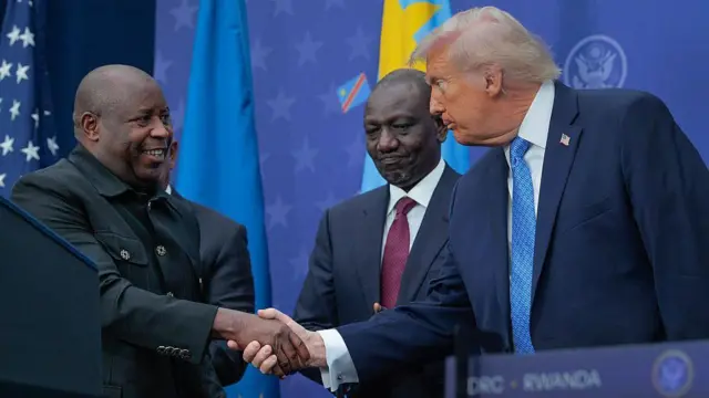 Perezida w'u Burundi Évariste Ndayishimiye, arimo kumwenyura, ahanye ikiganza na Perezida w'Amerika Donald Trump. Iruhande yabo hagati hari Perezida wa Kenya William Ruto. Perezida Ndayishimiye yambaye ikoti ry'umukara rikoze nk'ishati, naho Perezida Trump yambaye ikositimu y'ubururu bwijimye, ishati y'umweru na karuvati irimo utubara duto tw'ubururu. Ifoto yafotorewe i Washington DC, muri Amerika, ku itariki ya 4 Ukuboza (12) mu mwaka wa 2025, ubwo Perezida w'u Rwanda na Perezida wa Repubulika ya Demokarasi ya Congo bashyiraga umukono ku masezerano y'amahoro.