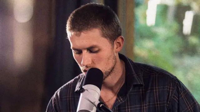 Un hombre de pelo castaño corto canta ante un micrófono. Lleva una camisa a cuadros.