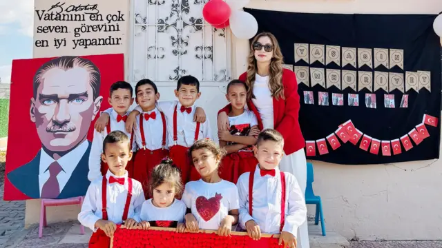Kübra Tüm ve öğrencileri bayramda poz veriyor. Hepsi kırmızı beyaz giyinmiş, öndeki çocukların elinde bir Türk bayrağı var. Yanlarında kırmızı arka plan üzerine bir Atatürk protresi duruyor.
