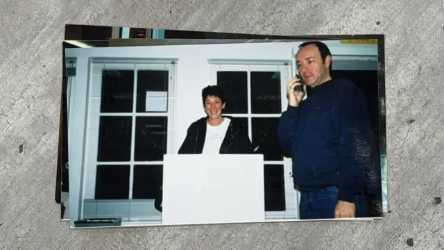 A woman, Ghislane Maxwell, with a short haircut smiles and stands behind a white podium to her left a man, Kevin Spacey, has a cell phone on his right ear. He is wearing a navy shirt and blue jeans.