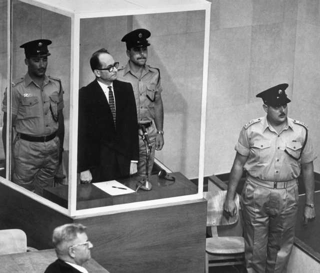 Adolf Eichmann vestido de traje y corbata con dos guardias a los lados, dentro de un cubilo protegido por vidrios