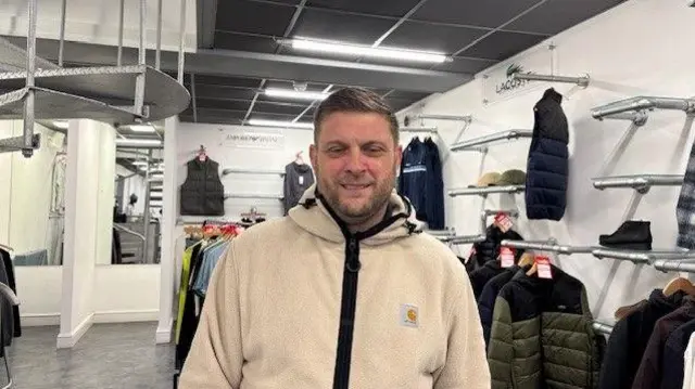Un hombre que viste una chaqueta de color crema se encuentra en una tienda con percheros con ropa de hombre al fondo.