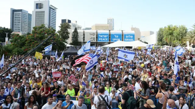 Miles de personas reunidas en un gran espacio abierto, agitando banderas de Israel y una de Estados Unidos. Al fondo, unos carteles piden "paz en Israel". 