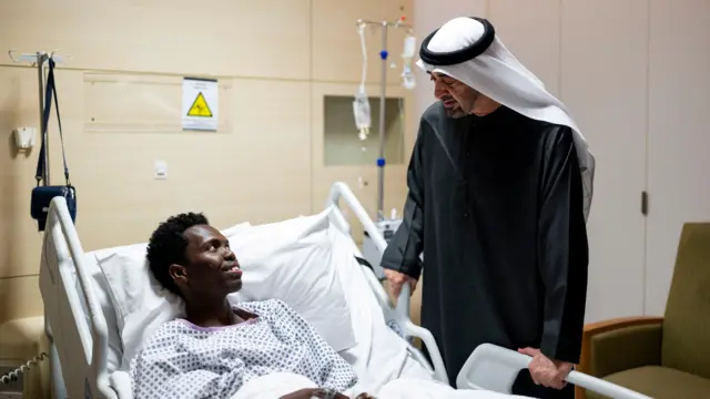 رئيس دولة الإمارات العربية المتحدة الشيخ محمد بن زايد آل نهيان يرتدي جلباباً أسود وعقالاً أبيض، داخل أحد المستشفيات بأبو ظبي حيث يزور شخصاً أُصيب، جراء الهجمات الإيرانية، ويتلقى العلاج في المستشفى. ويستلقي الشخص المصاب على سرير.