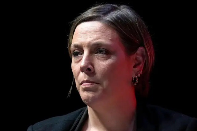 Retrato de Jess Phillips em um palco ou evento com fundo escuro. Ela veste um casaco escuro e usa vários brincos