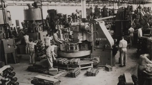 A imagem em preto e branco mostra o interior de uma fábrica com diversos operários trabalhando ao redor de grandes máquinas industriais. Os trabalhadores usam macacões claros e manipulam peças metálicas espalhadas pelo chão e sobre bancadas, enquanto equipamentos de grande porte ocupam quase todo o espaço do galpão.