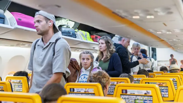 Utasok szállnak fel egy Ryanair járatra: a fedélzeten nyitva vannak a csomagtartók, néhányan már ülnek, míg mások a folyosón sorban állnak.