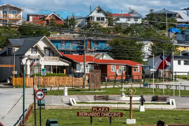 Plaza Bernardo Ohiggins in Puerto Williams on a bright day with clouds in the blue sky. Multi-coloured houses are at the end of a street with grassy borders. Blue houses are behind them, on a hillside and higher up the slope are white and brown houses. A sign saying "Plaza Bernando Ohiggins" is in the foreground.