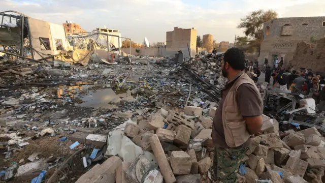 رجل يرتدي بنطالاً يشبه البنطال العسكري وقميص بني اللون، من بين أشخاص قرب أنقاض قاعة مناسبات تعرضت لضربات جوية أمريكية في صنعاء