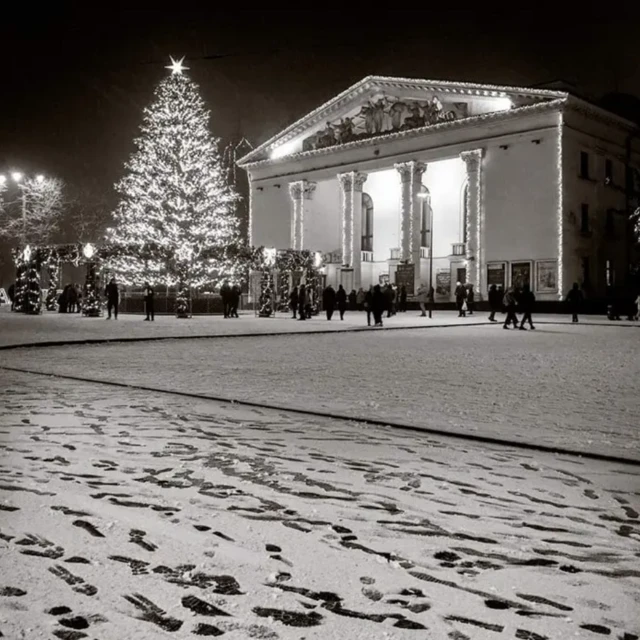 Заснеженная площадь, на которой стоит елка, а позади подсвеченный театр. На площади гуляют люди. 