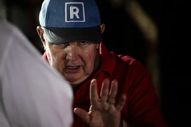 Rafael López Aliaga, con una gorra azul con el logo de su partido y la palma de la mano izquierda levantada a la altura de su pecho. 