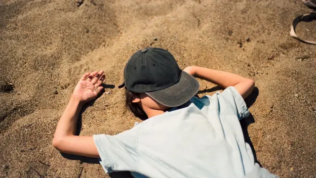 Дитина лежить на піску, обличчя закрите кепкою