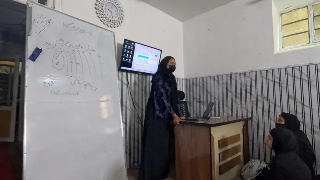 معلم که لباس بلند و ماسک صورت سیاه دارد، پشت میز ایستاده است. پشت سر او کامپیوتر به دیوار نصب شده و در پهلویش تخته سفید است. 