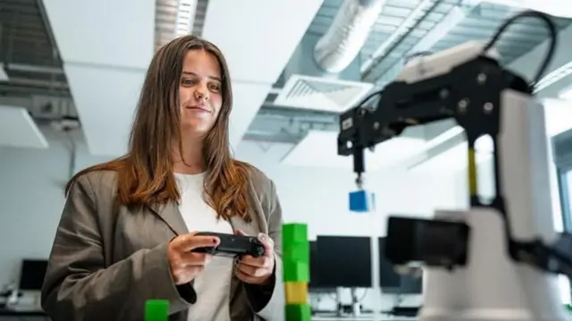 Una mujer con cabello castaño largo, una blusa blanca y una chaqueta marrón sosteniendo un pequeño dispositivo negro en una habitación blanca con dispositivos mecánicos en su interior.