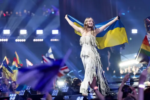 На сцені великого концертного залу людина в світлому костюмі з бахромою стоїть із розгорнутим українським прапором над головою. На передньому плані глядачі махають різнокольоровими прапорами, а позаду видно яскраві сценічні вогні та затемнений зал.