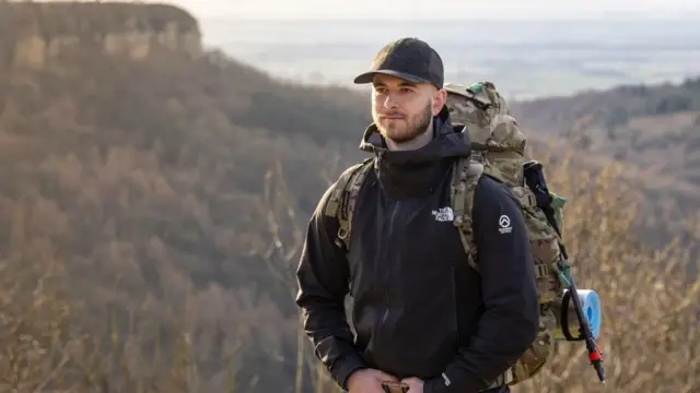 Un hombre se encuentra en un terreno rural. Al fondo se ven colinas. Lleva una chaqueta negra, una gorra de béisbol y una mochila grande de estilo militar.