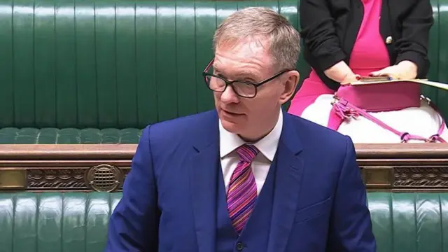 イギリス下院の緑色のベンチを背に、青い三つ揃いスーツに紫や青の斜め縞のネクタイをしたブライアント氏が答弁している