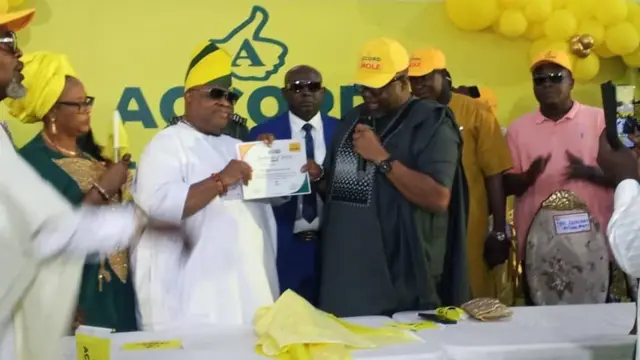 Gómìnà Ademola Adeleke wọ aṣọ funfun, oun ati awi ẹni to wọ aṣọ dudu mu iwe éri kan dani. Awọn eeyan to wa nibẹ n wo wọn