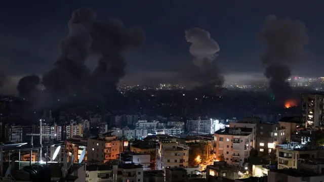 夜の都市で、数カ所から上空に煙が上っている。手前には建物群が見える