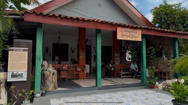 Sebuah rumah tua dengan tiang berwarna hijau. Ada beberapa orang duduk-duduk di teras. Ada pohon natal menjelang perayaan Hari Natal. Ada tulisan: "Living Museum, Roemah Toegoe" dan papan penjelasan.