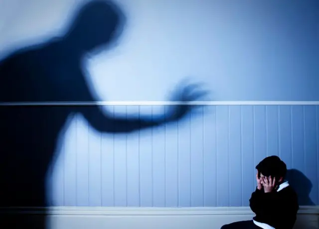 Bir yetişkin gölgesi köşeye sıkışmış ve öfkeyle yüzünü kapamış bir çocuğu eli havada korkutuyor. 