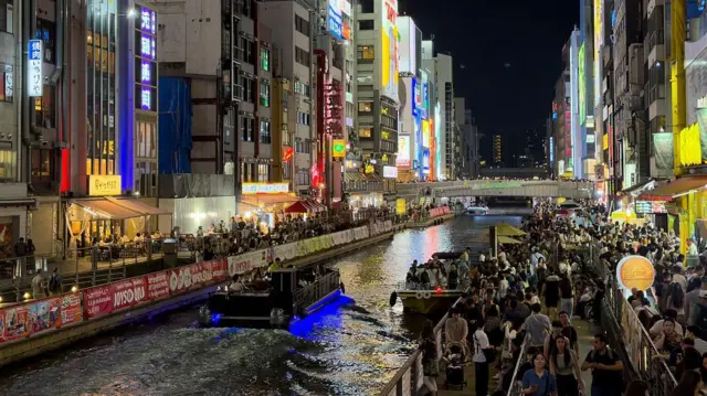 Japonya'nın Osaka kentindeki Dotonbori Kanalı boyunca akşam kalabalıkları. Işıklı reklam panoları, restoranlar ve gezi tekneleri, Namba eğlence bölgesindeki en tanınmış gece hayatı manzaralarından birini oluşturuyor.