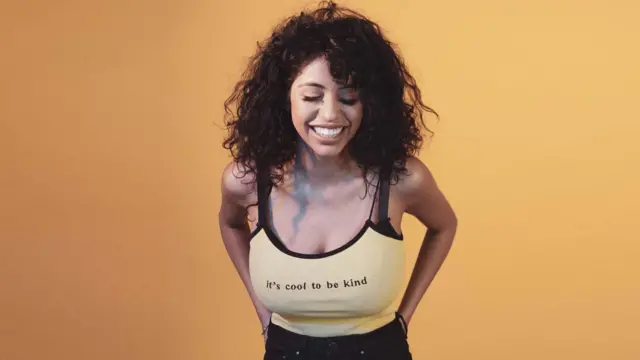 Ranvia smiling in front of an orange background, where she is stood wearing a strappy vest top which says 'it's cool to be kind.' The photo was taken before her breast reduction surgery
