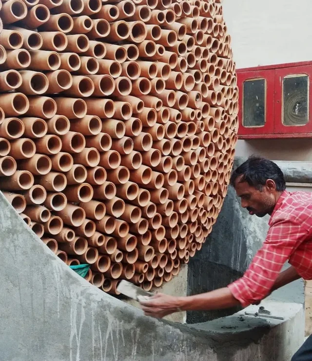 La surface de plusieurs rouleaux d'argile empilés en forme de ruche est finalisée par un homme brun en chemise rouge.