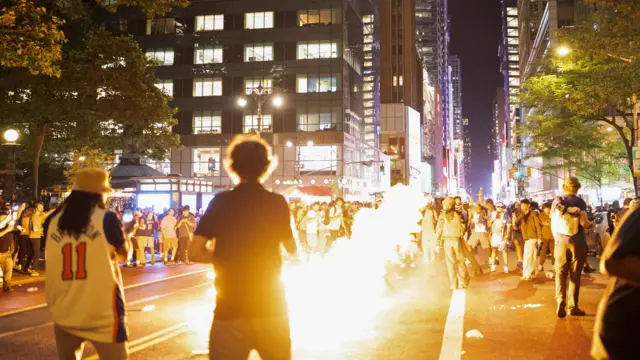 A man walks away from the camera toward a browd on a New York street - there is fire in front of him