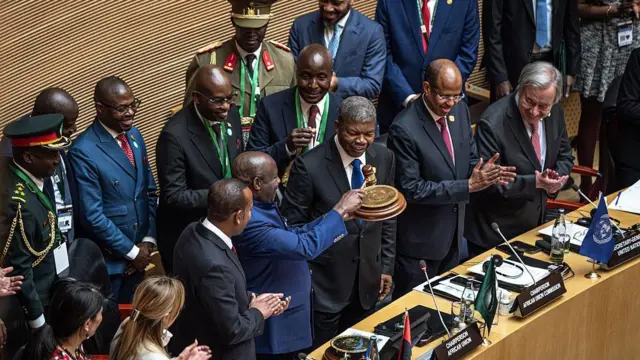 Perezida w'Angola João Lourenço (hagati) ahererekanya ububasha na Perezida mushya wa AU akaba na Perezida w'u Burundi, Évariste Ndayishimiye, mu nama isanzwe ya 39 y'inteko ya AU, ku cyicaro cya AU, i Addis Ababa muri Ethiopia, ku wa gatandatu, ku itariki ya 14 Gashyantare (2) mu 2026. Bari imbere, bakikijwe n'abategetsi benshi, bose bahagaze. Bamwe barimo gukoma amashyi. Imbere yabo hari ameza ariho indangururamajwi n'amabendera mato, arimo n'irya AU na UN. 