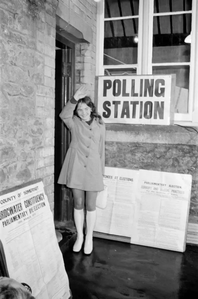 Черно-белая фотография с избирательного участка, юная девушка машет рукой.