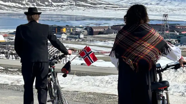 Norwegowie prowadzący rowery podczas swojego święta narodowego na Svalbardzie, z wodą i szczytami lodowców w tle.