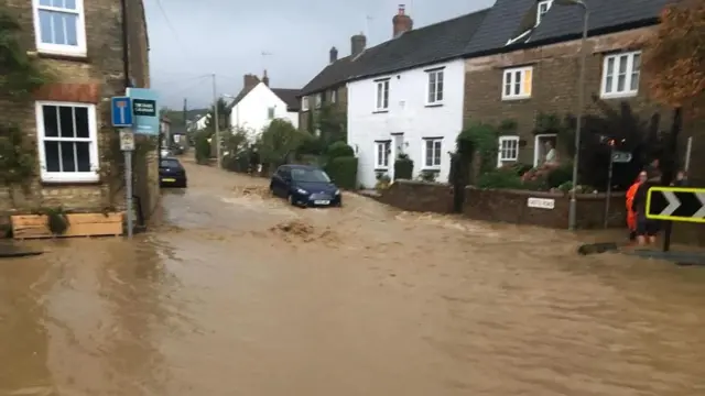 Una calle residencial está gravemente inundada, con agua fangosa fluyendo entre hileras de casas. Varios coches están parcialmente sumergidos, y una persona con ropa de alta visibilidad permanece cerca del arcén mientras el agua de la inundación se extiende por toda la calzada.