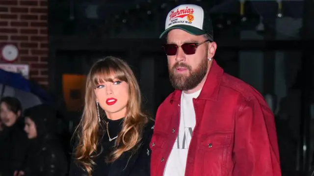 Taylor Swift ve Travis Kelce yan yana yürürken, üstlerine flaş patlamış. Taylor'ın üstünde siyah bir kıyafet var, Travis ise beyaz tişörtünün üstüne kırmızı bir gömlek giyip gözlük ve şapka takmış.