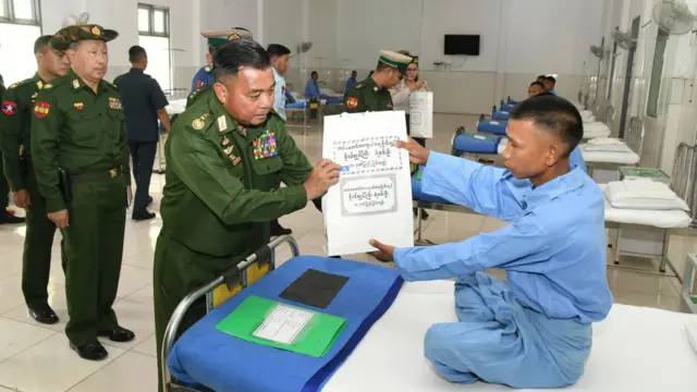 ဗိုလ်ချုပ်ကြီးရဲဝင်းဦးက စစ်ဆေးရုံက လူနာတဦးကို လက်ဆောင်ပေးနေတာဖြစ်ပြီး သူ့နောက်မှာ ဗိုလ်မှူးချုပ်ဇော်မျိုးဟန်က သတိစွဲပြီး ရပ်နေပါတယ်။