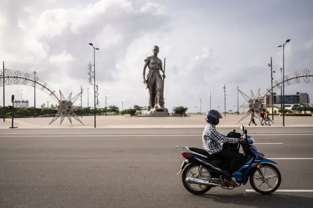 Un automobiliste sur une moto bleue et noire, vêtu d'une chemise à carreaux, passe devant une grande statue d'Amazone en arrière-plan.