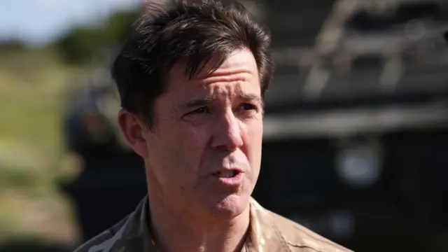 Major General Tom Bewick, who is middle-aged and has short brown hair, is seen squinting in the sun as he speaks to journalists at RAF Akrotiri 