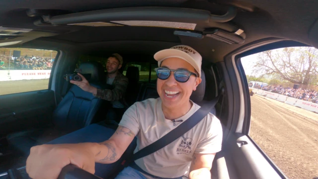 Ash Pérez aparece dirigindo um carro. No banco de trás há um homem com uma câmera.