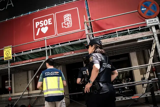 Policías nacionales custodian la entrada de un edificio en obras con carteles del PSOE, bajo un andamio y señales de seguridad.