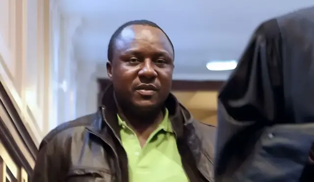 Claude Muhayimana yambaye ijaketi y'umukara n'ishati y'icyatsi kibisi