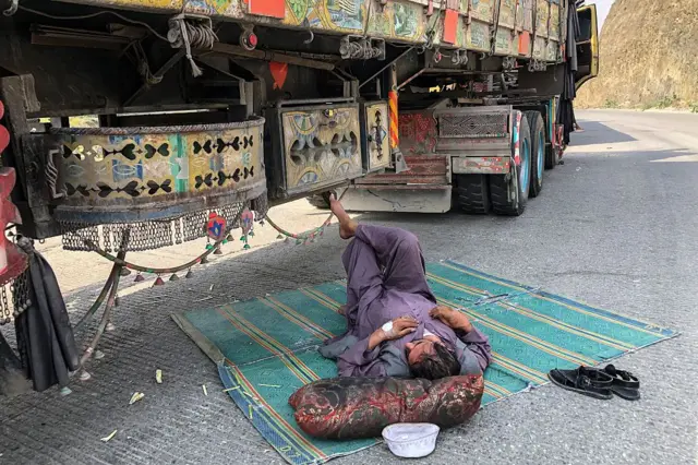 یک راننده در مرز افغانستان و پاکستان زیر سایه موتر لاری خود روی یک فرش سبز رنگ خوابیده است