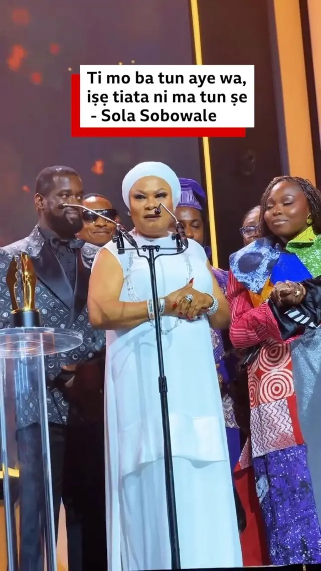 Sola Sobowale wọ asọ kaba gbagẹrẹ alawọ funfun, o duro siwaju ẹrọ amohunbugbẹmu, awọn eeyan kan to yi ka