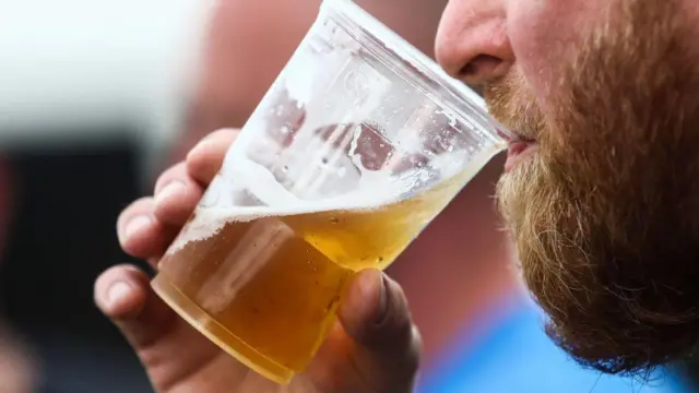 Close-up de um homem barbado bebendo cerveja em um copo plástico, com a bebida dourada e a espuma em destaque.