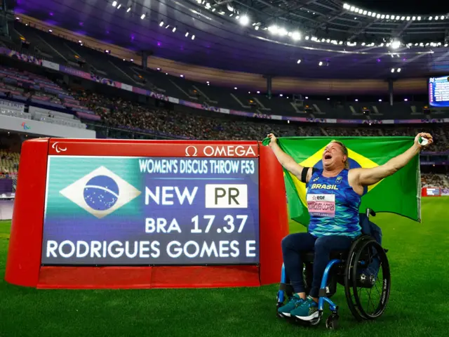 Elizabeth Gomes celebra su récord paralímpico al costado de una pantalla que muestra su logro mientras sostiene con sus manos la bandera de Brasil.