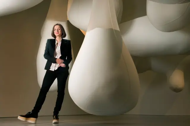 Sarah Thornton, vestida con una chaqueta y un pantalón negros y una blusa blanca, al lado de una obra de arte que consiste en formas gigantes de color blanco