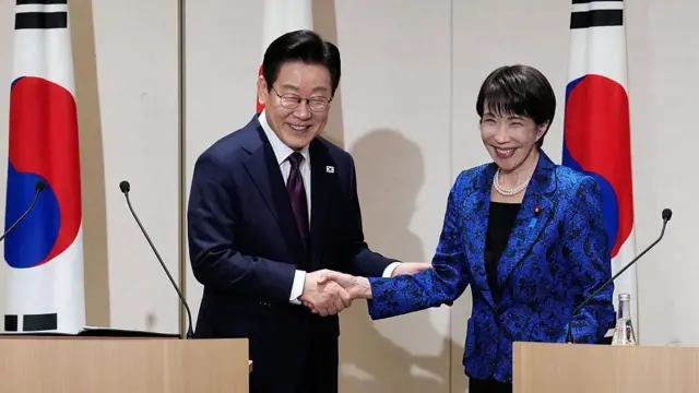 ダークスーツ姿の李氏と、青いジャケットを着た高市氏が、演壇で握手している。背後には両国の国旗が立てられている