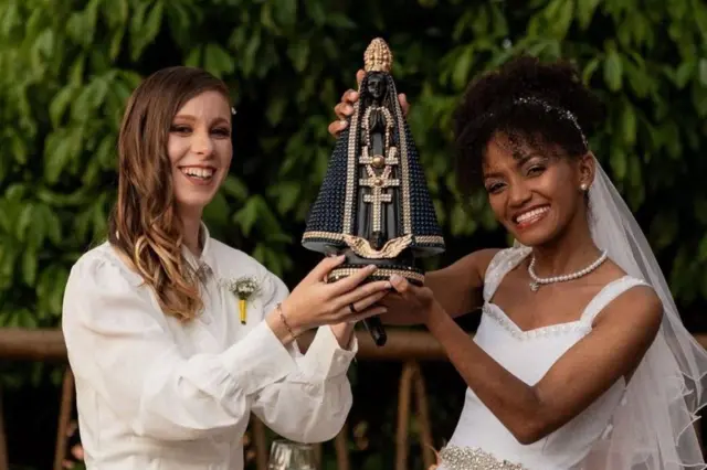 Fotografia colorida mostra duas noivas sorrindo e segurando juntas uma estátua de Nossa Senhora Aparecida. A mulher da esquerda, loira, usa um camisa branca com uma flor na lapela. A mulher da direita, negra, usa um vestido de noiva