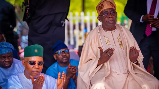 Nigeria President Bola Tinubu wear creamy-brown attire, im sitdon for chair beside Vice President Kasim Shettima wey wear greencap and sky blue cloth with dark sunglasses.