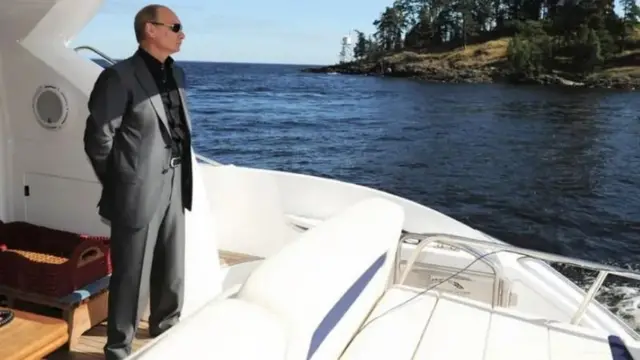 Putin ari mu bwato yambaye imyenda isa i pantaro n'i kote bifite ibara ry'ivu n'ishati y'umukara. Yambaye n'amadarubini