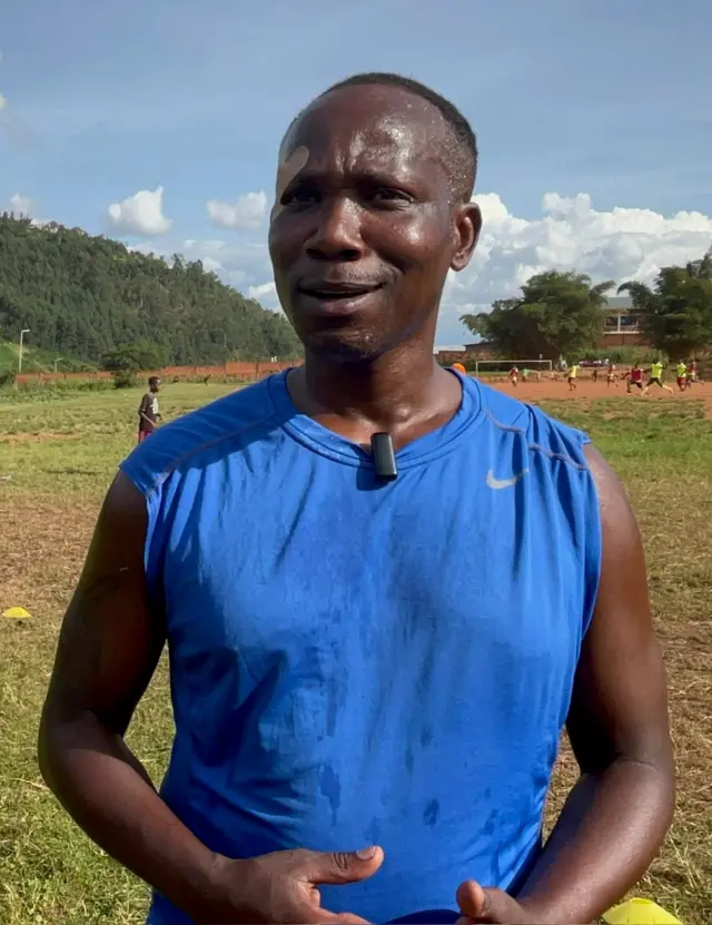 Fabrice Twagizimana yambaye umupira w'ubururu udafite amaboko, akoze ikimenyetso n'ibiganza, arimo kuvuga areba muri 'camera'. Yabize icyuya mu mutwe, ku ijosi, ku maboko, mu maso no mu gatuza. Ahagaze mu kibuga, inyuma ye kure hari abakinnyi barimo gukina umupira w'amaguru, hirya hari ishyamba n'inzu. 